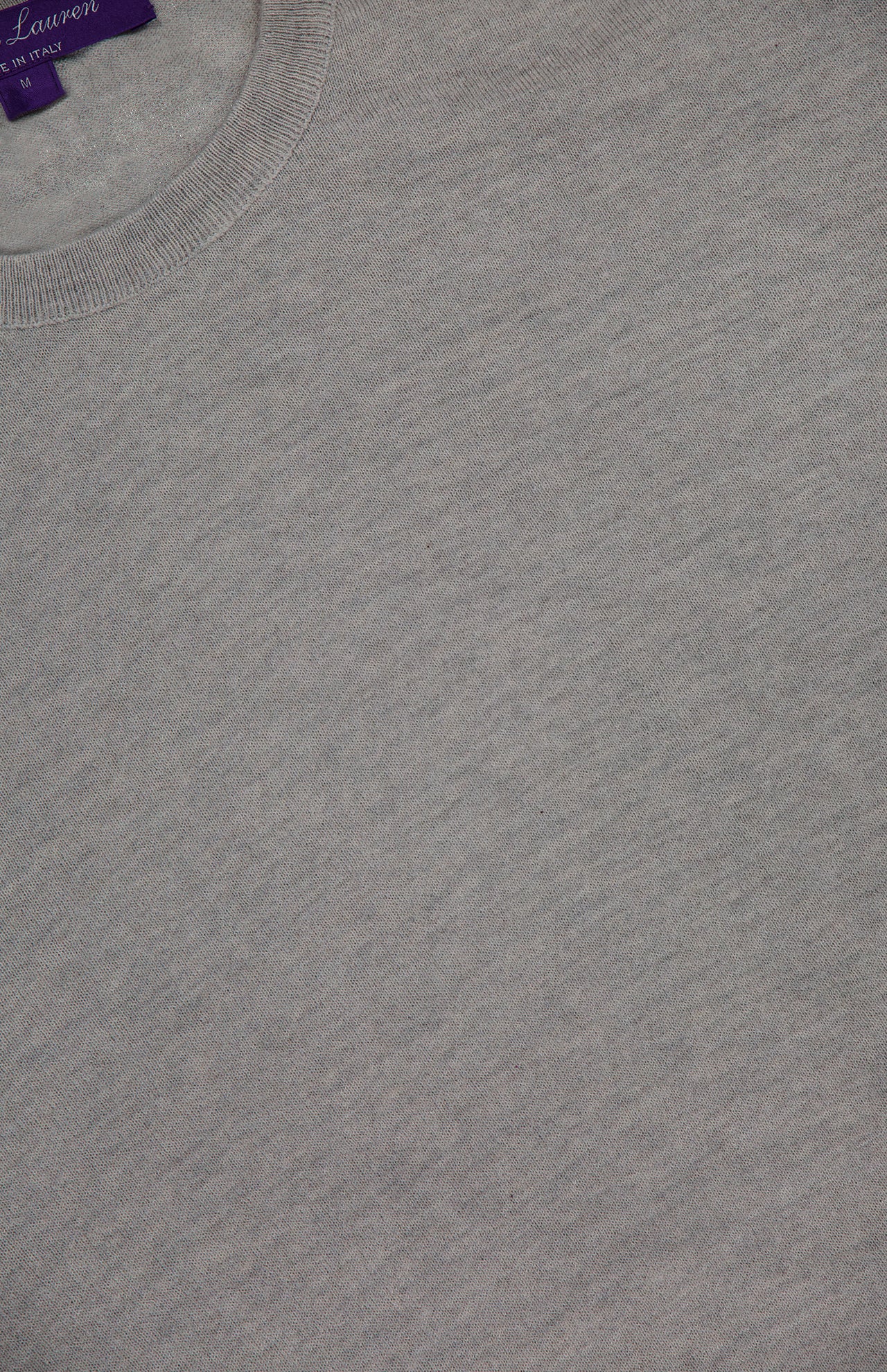 Close-up view of the light gray Cashmere Crewneck Pullover Sweater from RALPH LAUREN PURPLE LABEL, featuring a ribbed collar. A partially visible purple tag in the top left corner indicates it is made in Italy. The image highlights the fabric texture and detailed stitching.