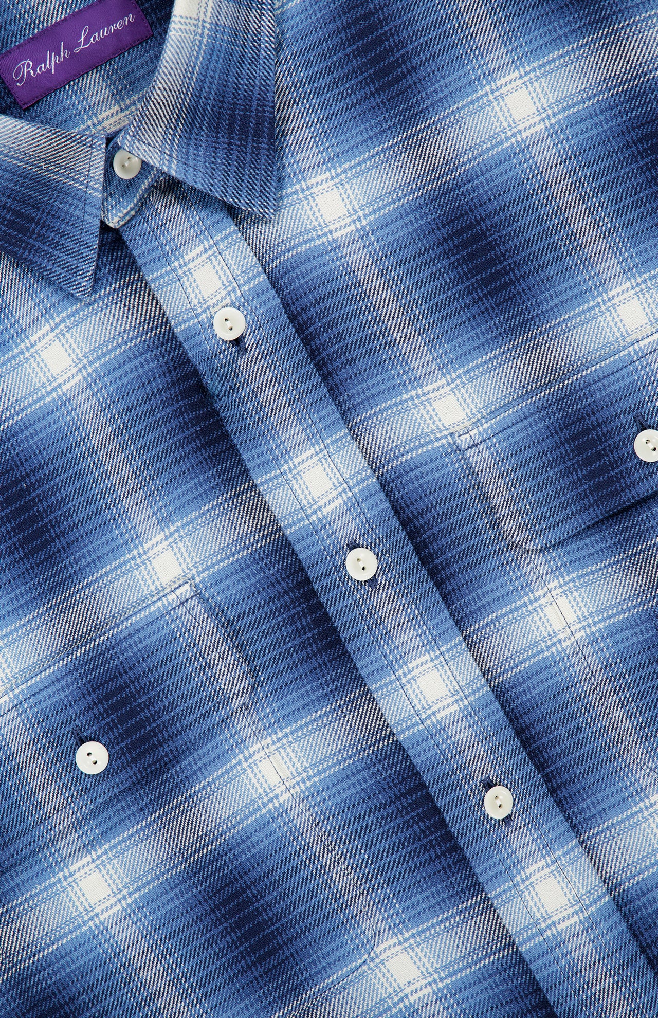 A close-up of the Cooper Cotton Plaid Sport Shirt highlights its blue and white plaid button-up design with white buttons and a collar. The shirt features a purple RALPH LAUREN PURPLE LABEL at the top left, emphasizing this classic sport shirt from Ralph Lauren’s premium collection.