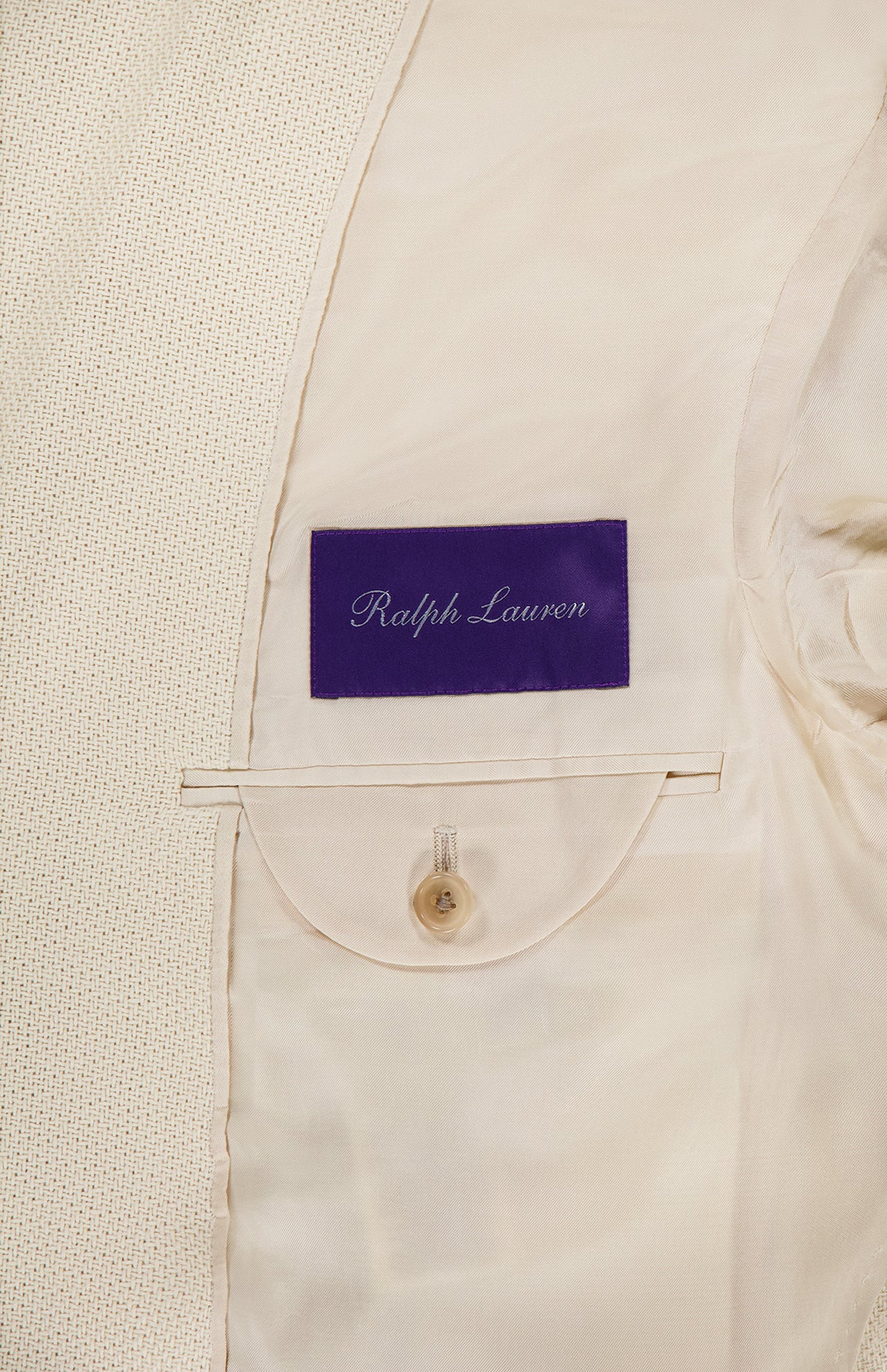 Close-up of the inside of a light beige Cotton Panama Weave Double Breasted Sportcoat by RALPH LAUREN PURPLE LABEL, made in Italy, showing the signature purple label above a buttoned pocket on silky lining.