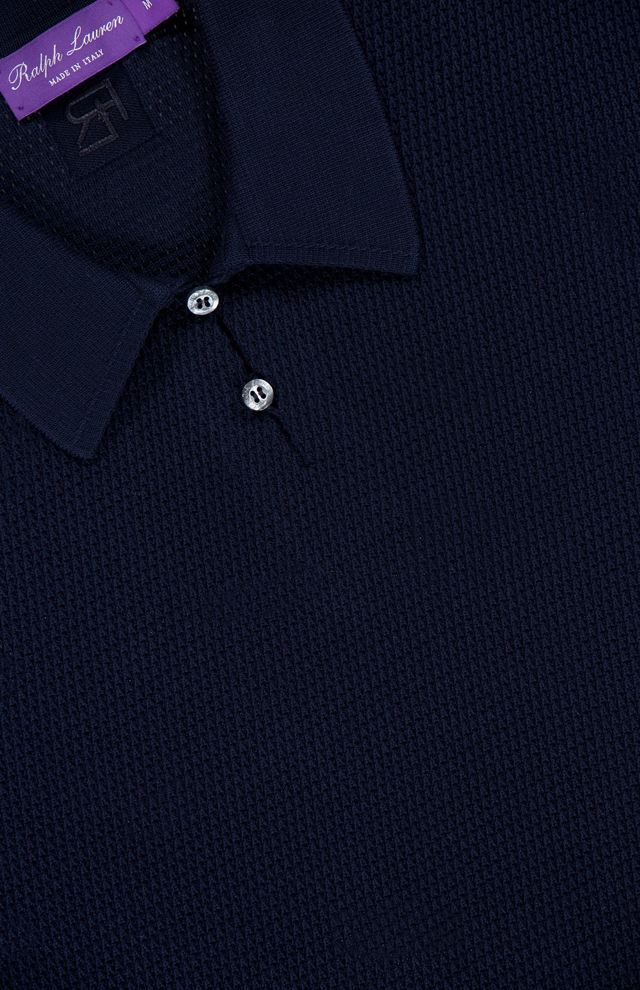 Close-up of a dark navy textured Cotton Short Sleeve Polo Pullover Sweater with two buttons and a collar, reflecting the refined craftsmanship characteristic of RALPH LAUREN PURPLE LABEL. A purple label inside the collar reads "Ralph Lauren Made in Italy" in white text, highlighting its luxurious origins.