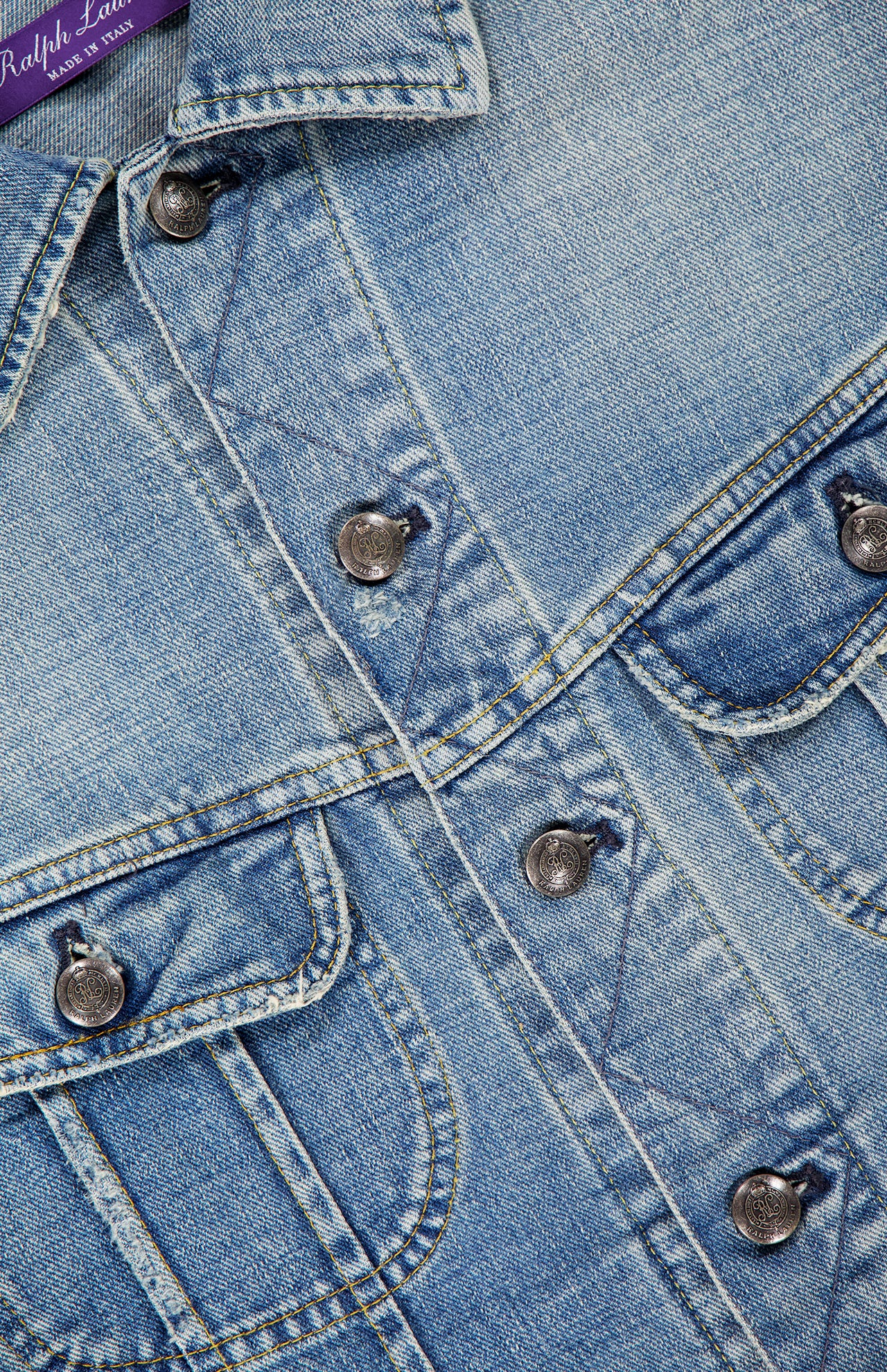 Close-up of the Lux Italian Denim Trucker Jacket from RALPH LAUREN PURPLE LABEL, featuring light blue Japanese denim with metal buttons, chest pockets, and prominent stitching details. A glimpse of the signature purple “RALPH LAUREN PURPLE LABEL” tag appears at the top left corner, accentuating the jacket’s subtle vintage fade.