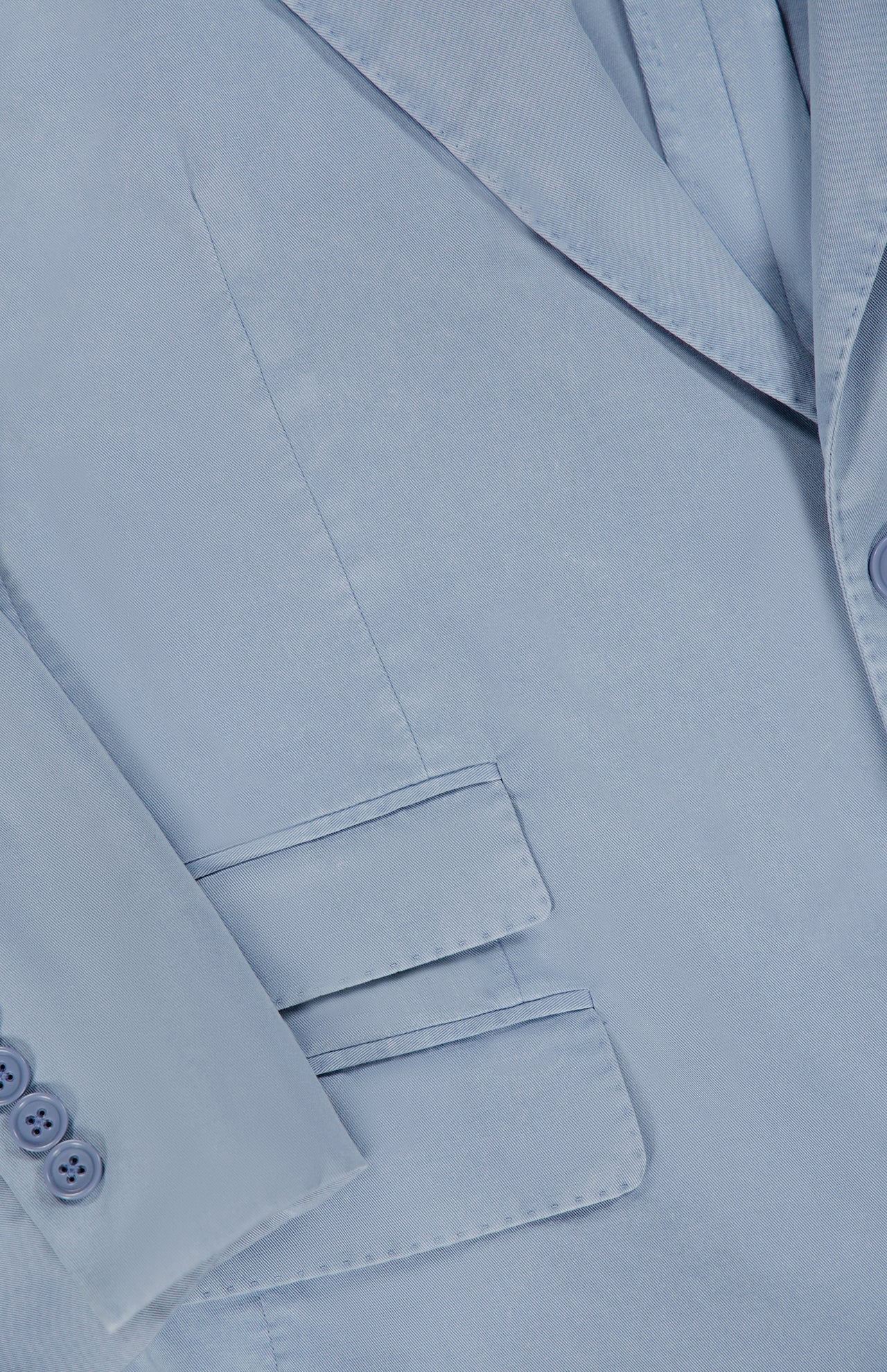 Close-up of the Garment Dyed Cotton Single Breasted Sportcoat by RALPH LAUREN PURPLE LABEL, featuring a light blue hue, a detailed lapel, a sleeve finished with three buttons, and two front flap pockets—all showcasing its vintage appeal and exquisite hand-tailored Italian craftsmanship.