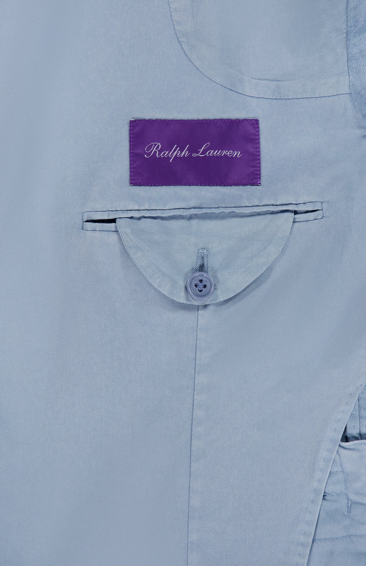 Close-up of the inner pocket of the Garment Dyed Cotton Single Breasted Sportcoat by RALPH LAUREN PURPLE LABEL, showcasing a curved flap with a button and crafted from stretch chino fabric. Above the pocket, a purple label with “Ralph Lauren” in white script highlights the vintage character of this hand-tailored Italian jacket.