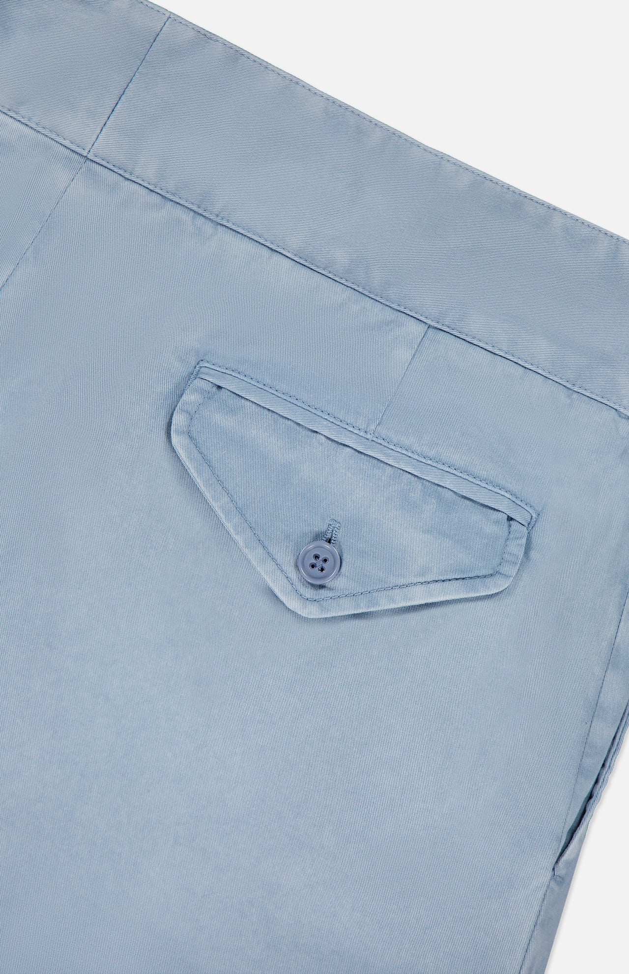 Close-up of a light blue, garment-dyed cotton fabric on the RALPH LAUREN PURPLE LABEL Garment Dyed Cotton Trouser Pant, showing a single back pocket with a triangular flap and matching blue button. The visible, precise stitching highlights the refined tailoring of these stretch chino trousers for enhanced comfort.