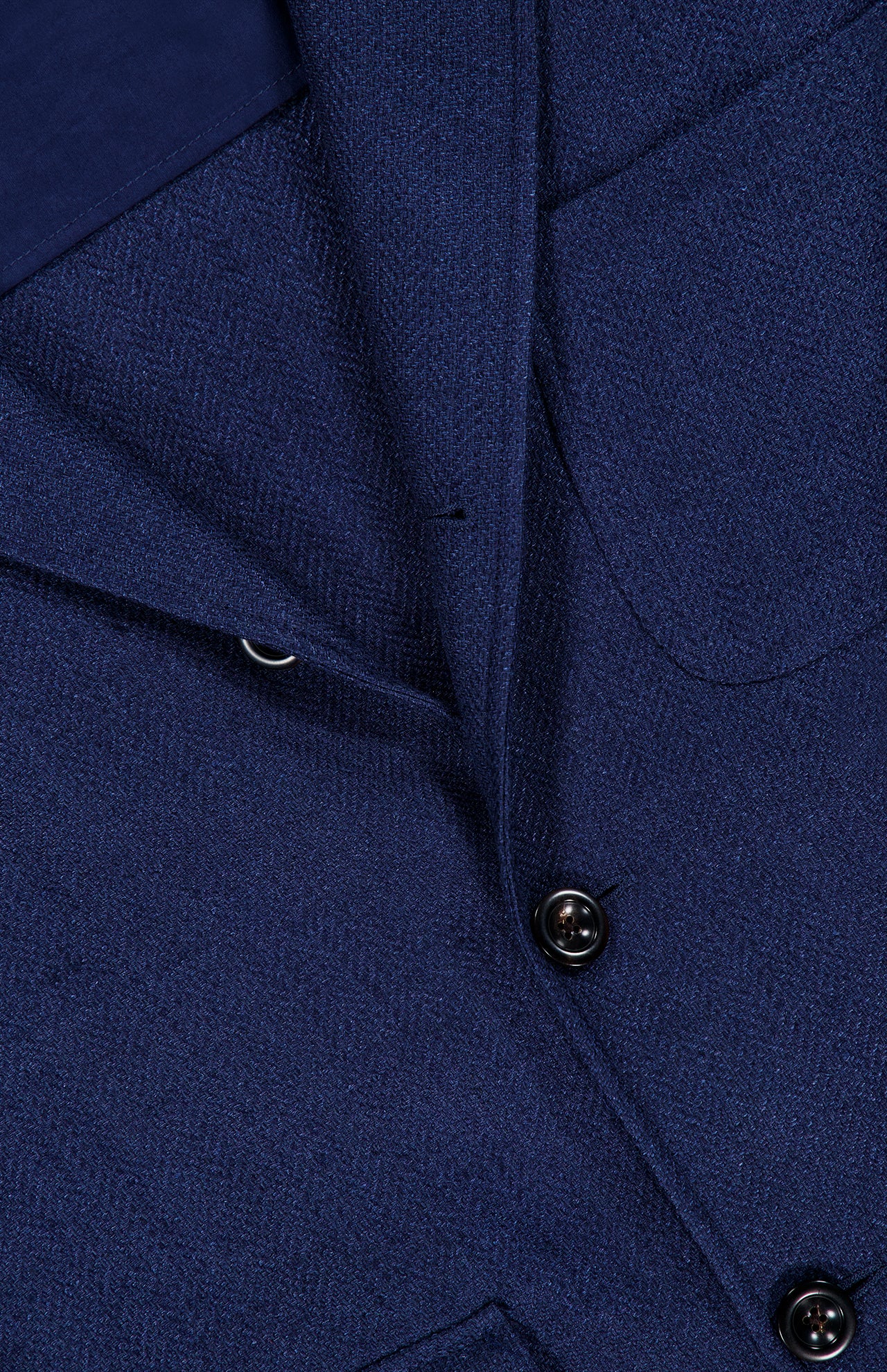 Close-up of the Herringbone Single Breasted Sportcoat by RALPH LAUREN PURPLE LABEL, featuring a blue textured fabric with distinctive stitching, buttons, and a front pocket. The image emphasizes the refined herringbone weave and deep indigo hue, capturing the handcrafted elegance for which Ralph Lauren Purple Label is renowned.