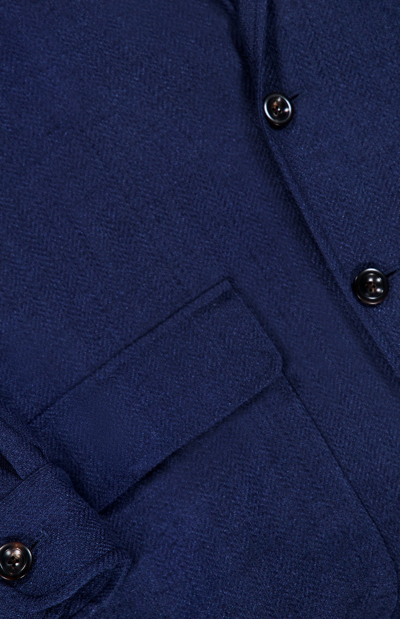 Close-up of the Herringbone Single Breasted Sportcoat by RALPH LAUREN PURPLE LABEL in dark blue, featuring a herringbone fabric, a flap pocket, sleeve cuff with button detail, and part of the front placket with two black buttons.