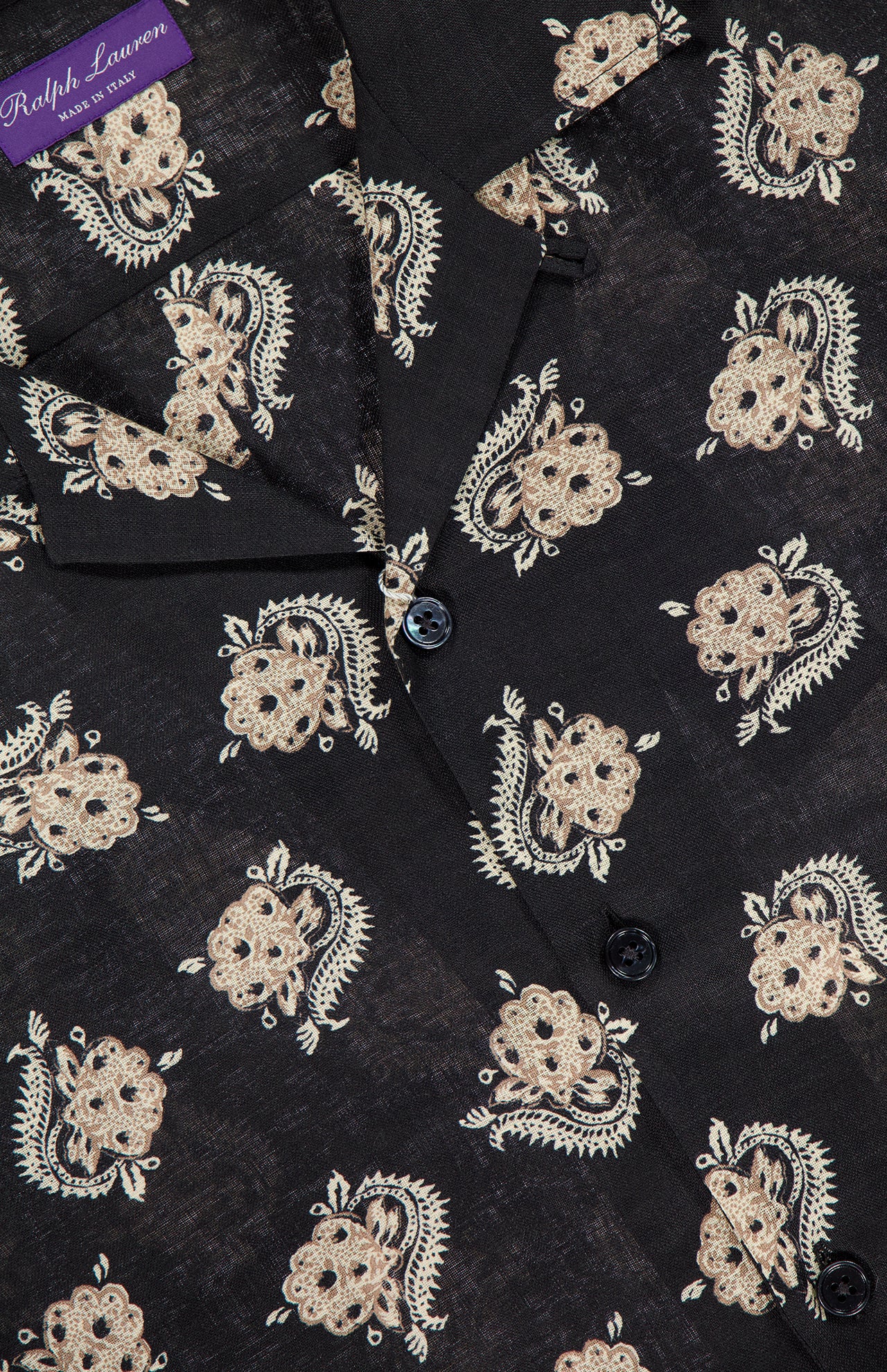 A close-up of a black RALPH LAUREN PURPLE LABEL Linen Print Short Sleeve Sport Shirt with beige floral and paisley designs, black buttons, and a visible purple brand tag on the collar.