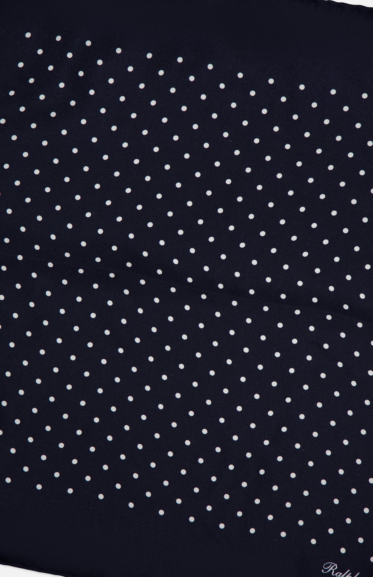 Crafted from luxurious silk, the Silk Foulard Dot Pocket Square by RALPH LAUREN PURPLE LABEL features a classic navy blue background adorned with evenly spaced small white polka dots. A visible crease adds character to the fabric, while "Ralph" appears partially in elegant white script at the bottom right corner—a refined signature of Purple Label craftsmanship. Made in Italy, this pocket square offers timeless style and exceptional quality.