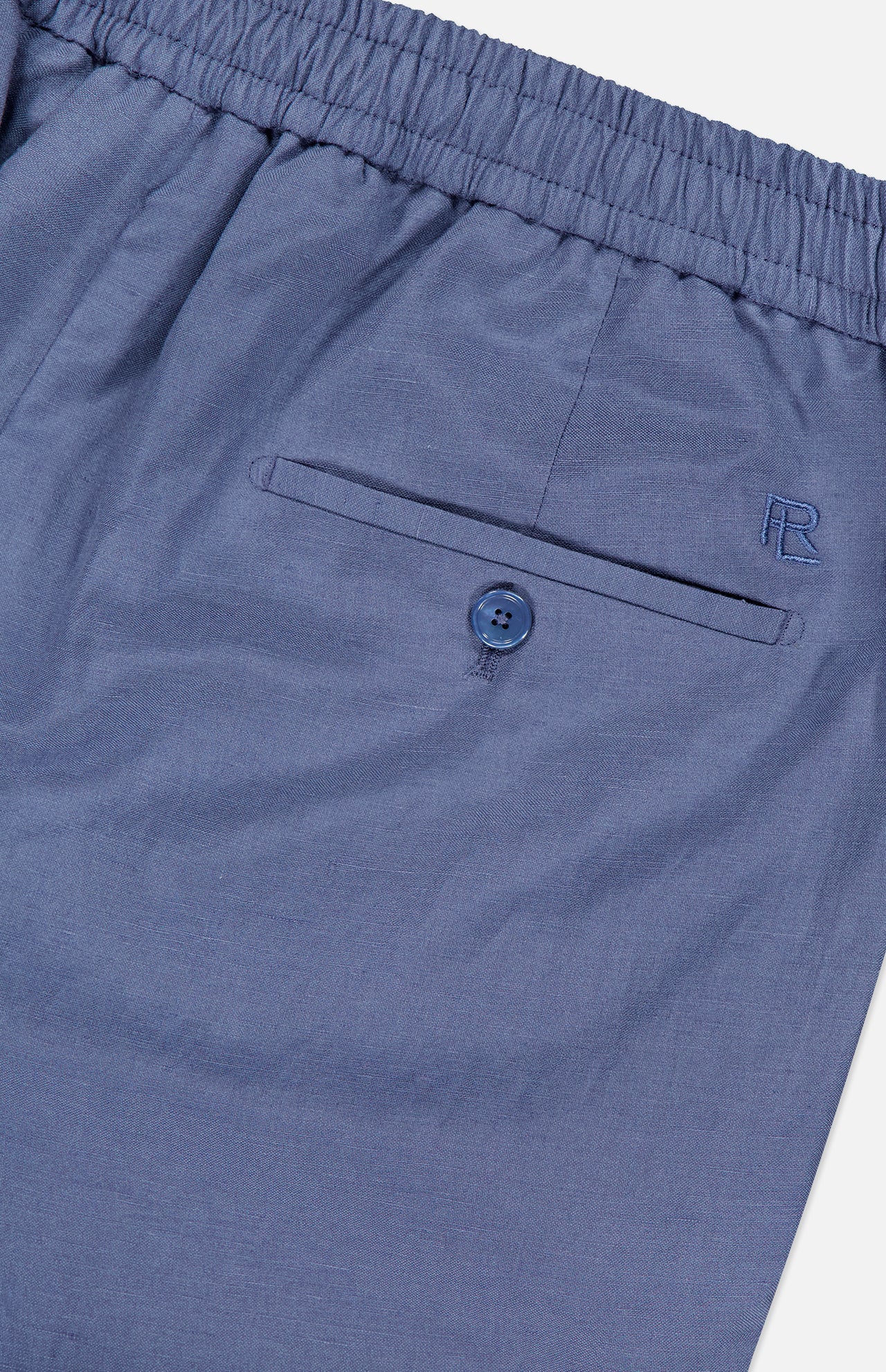 Close-up of the Silk Linen Dorset Flat Front Trouser by Ralph Lauren Purple Label, featuring blue silk-linen fabric, an elastic waistband, a buttoned back pocket, and an embroidered R logo.