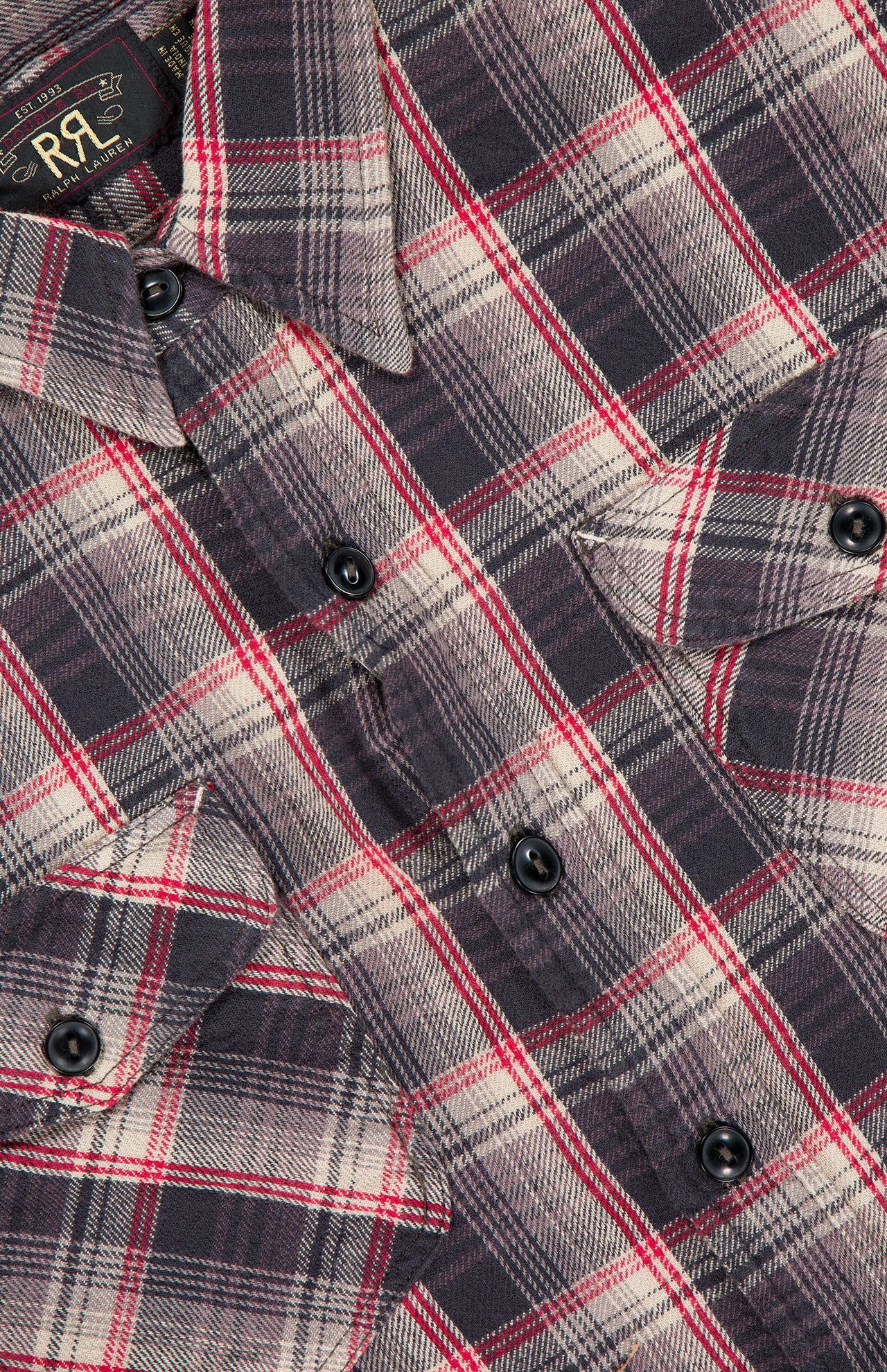 Close-up of the Jasper Cotton Long Sleeve Workshirt by RALPH LAUREN RRL, featuring a relaxed fit with black buttons, chest pockets, and a visible collar. The shirt displays a plaid pattern of red, black, and white stripes and includes a partially visible fabric label near the collar.