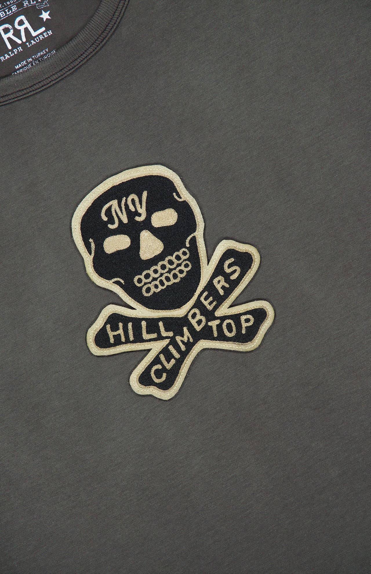 The Jersey Cotton Graphic Short Sleeve T-Shirt from RALPH LAUREN RRL is crafted from black cotton and features a bold motorcycle skull and crossbones patch with “NY Hill Climbers Top” emblazoned across it. The white and gold embroidered patch adds a subtle vintage touch to this standout design.