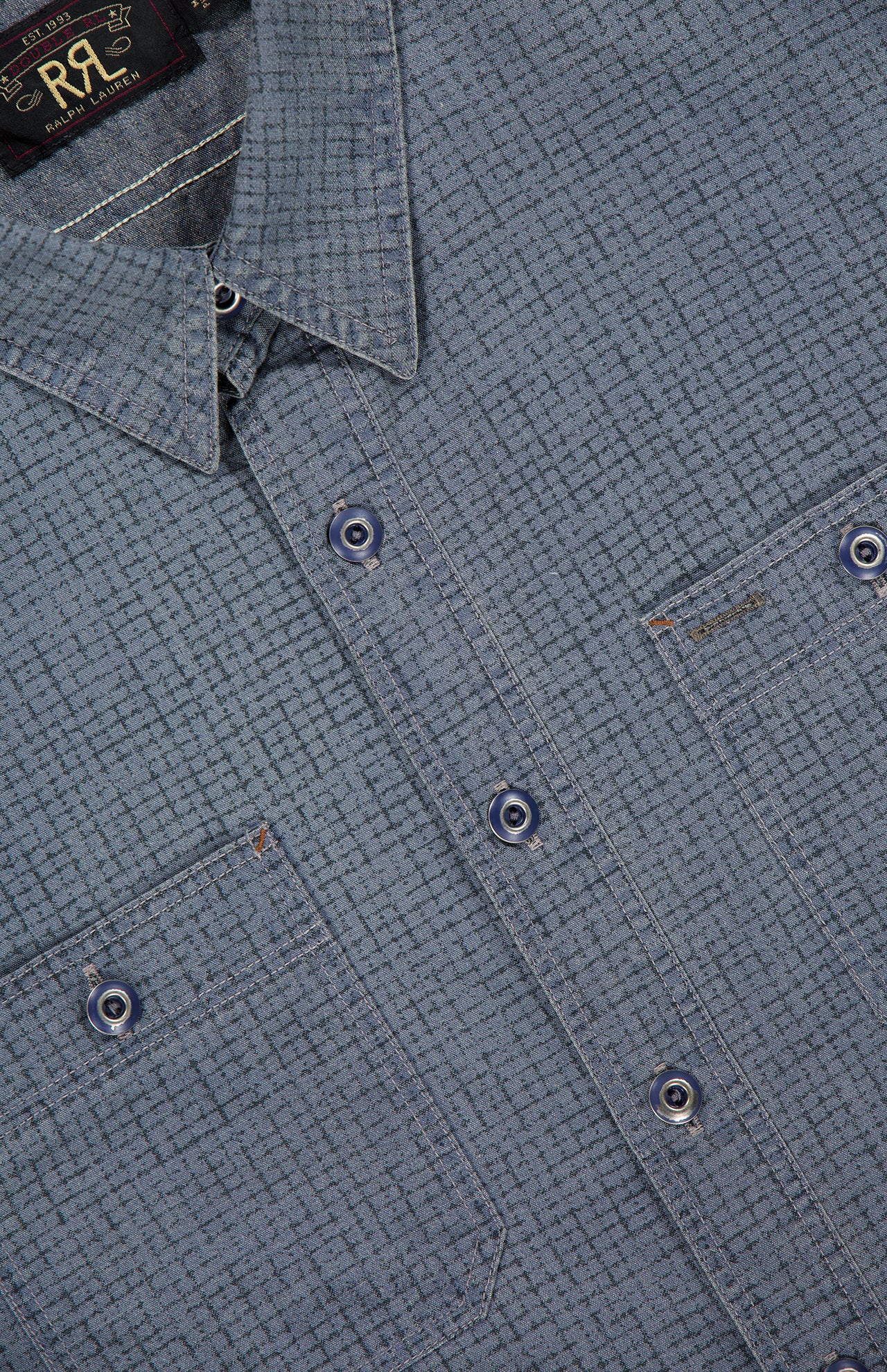 Close-up of the Montgomery Cotton Sport Shirt by RALPH LAUREN RRL, featuring blue textured cotton fabric, two front pockets, visible stitching, and a collar. A black label with gold text is partially visible near the neckline, emphasizing the classic workshirt fit.
