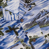 The RALPH LAUREN RRL Rayon 2-Pocket Camp Shirt features lightweight woven blue fabric, brown buttons, and tropical designs—mountains, palm trees, boats, water, and white flowers for a classic island vibe.