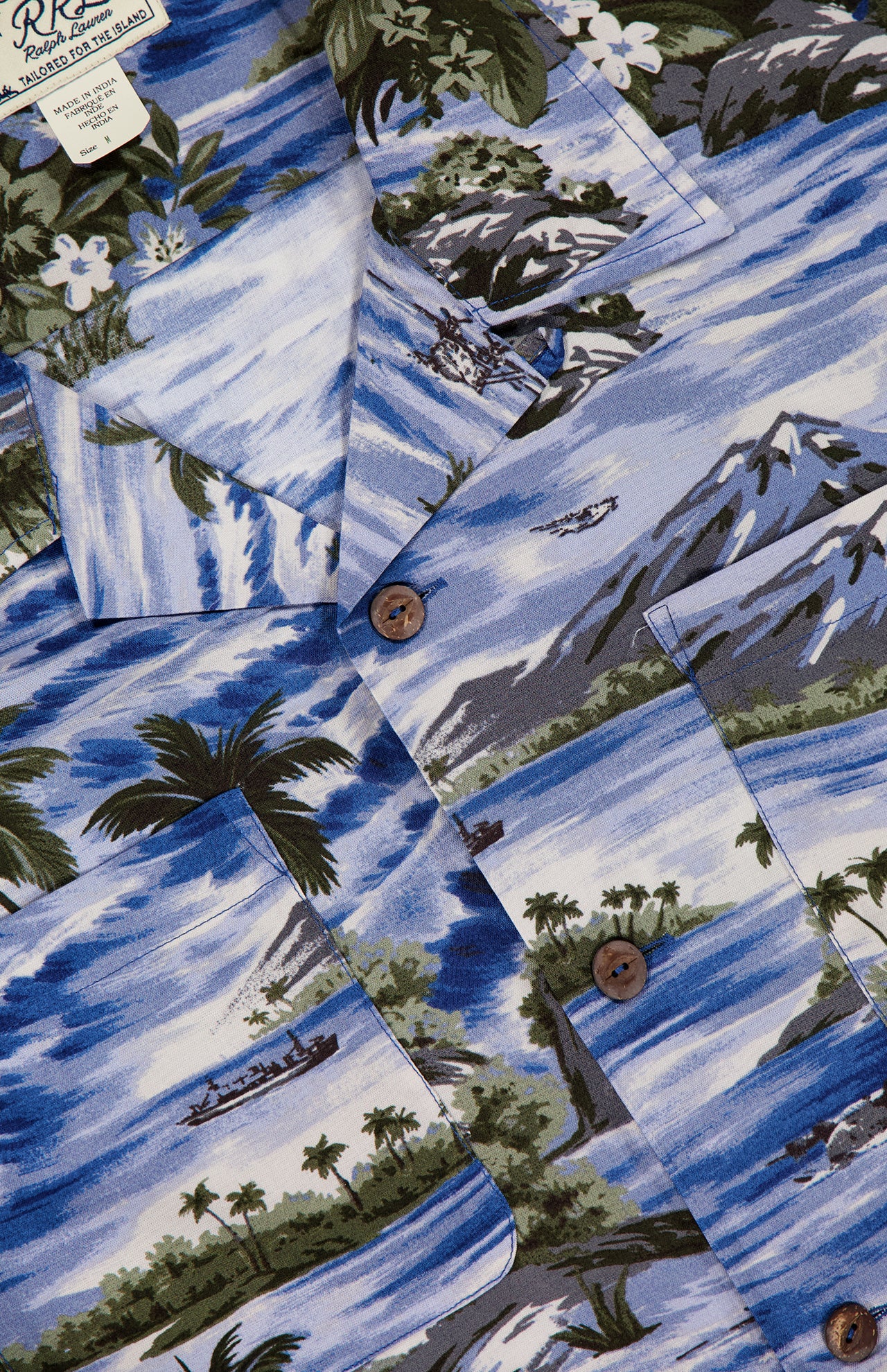 The RALPH LAUREN RRL Rayon 2-Pocket Camp Shirt features lightweight woven blue fabric, brown buttons, and tropical designs—mountains, palm trees, boats, water, and white flowers for a classic island vibe.