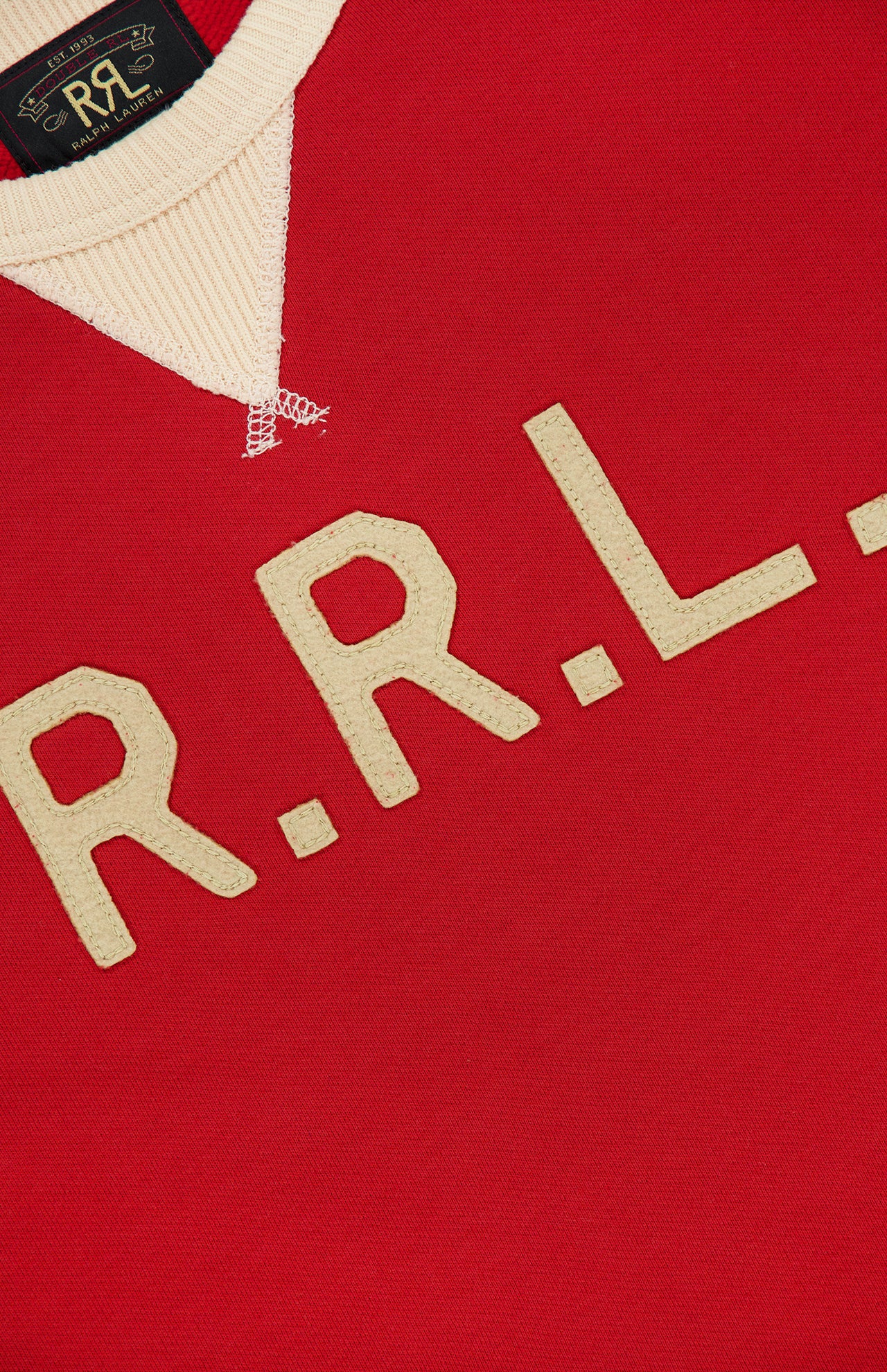 Close-up of the RALPH LAUREN RRL Terry Cotton Graphic Crewneck Sweatshirt, featuring a red hue with cream-colored “R.R.L.” lettering stitched on the front and a cream ribbed collar. Made from soft terry cotton, this pullover also showcases a black label with gold RRL branding near the neckline.