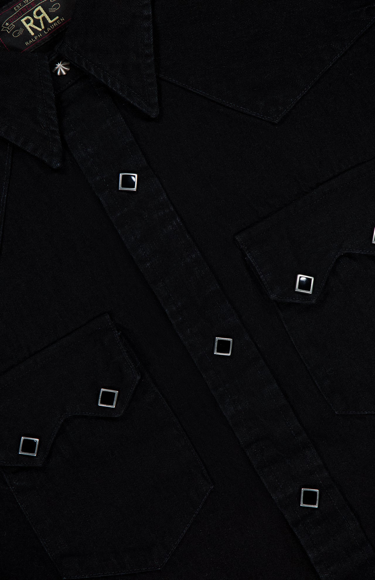 Close-up of the Western Denim Sawtooth Workshirt by RALPH LAUREN RRL, crafted from premium black Japanese denim. The shirt features two chest pockets with sawtooth flaps, silver square buttons, and visible contrast stitching. The collar and part of the RRL label are also visible at the top left.