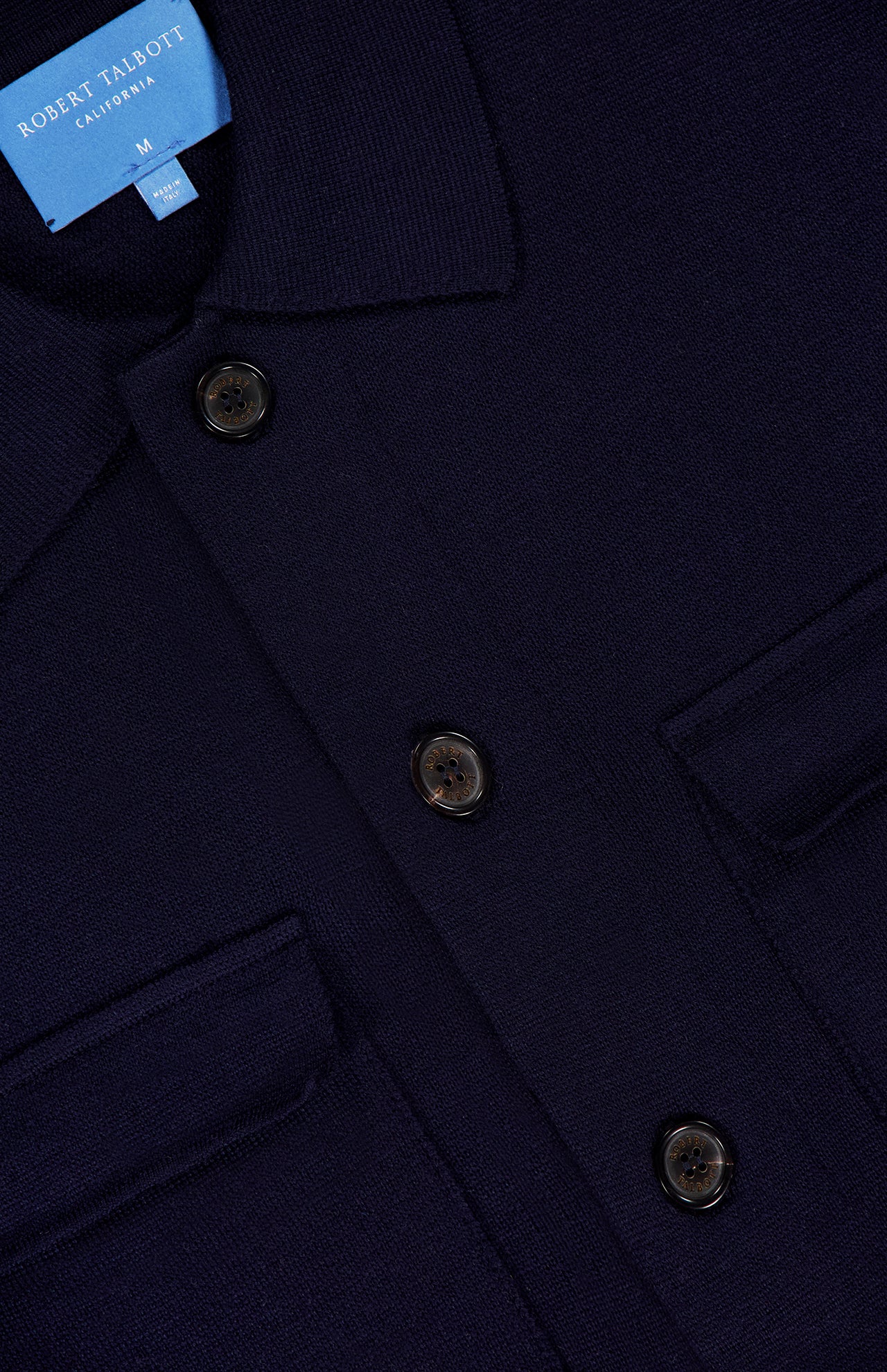 Close-up of the Landon Sweater Jacket by ROBERT TALBOTT, a navy blue button-up shirt-style jacket featuring a collar, two flap pockets, and a blue label reading Robert Talbott California. Crafted in a tailored fit, this hybrid sweater-jacket blends classic design with modern versatility.