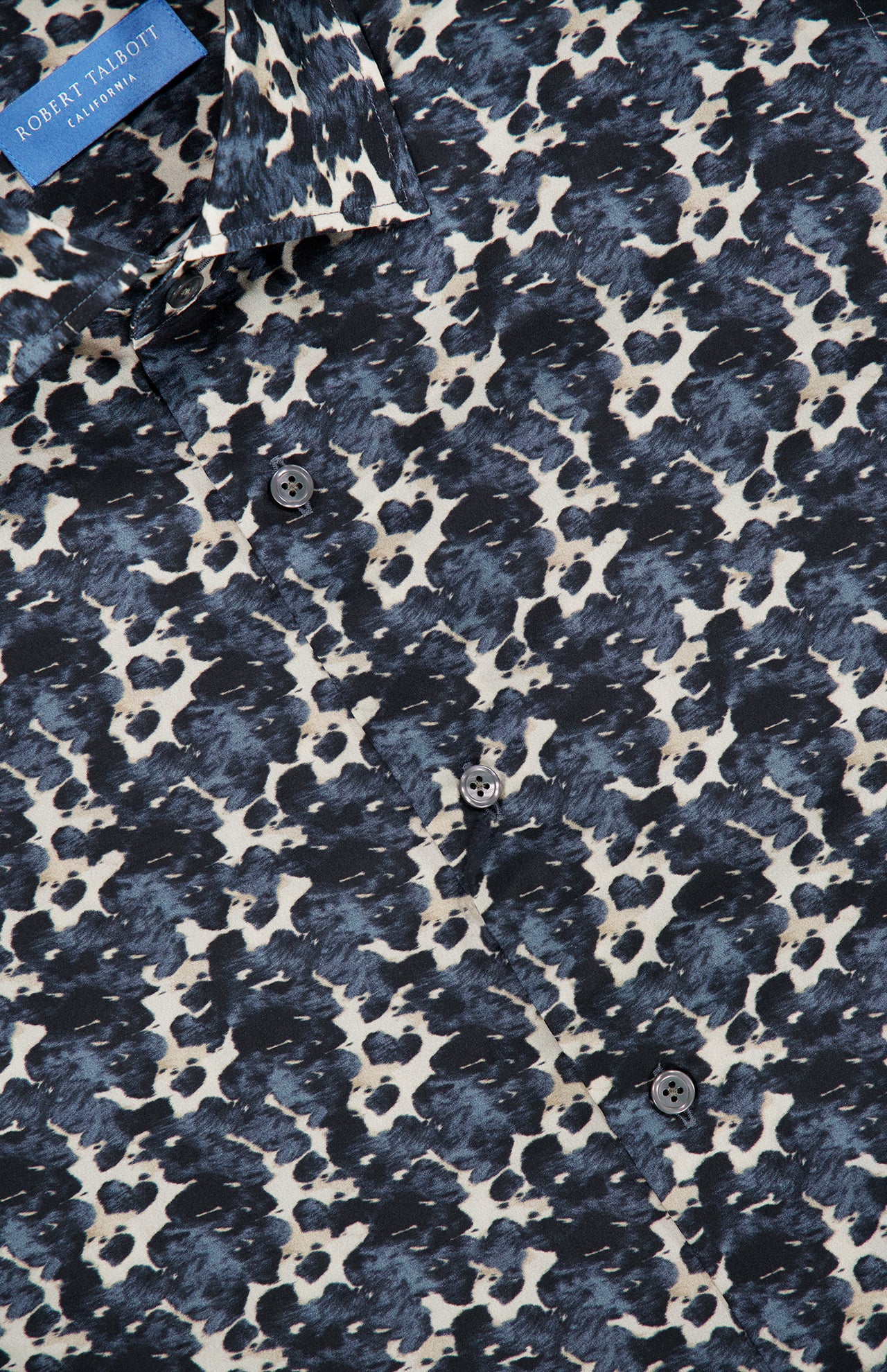Close-up of the Reynolds Printed Cow Hide Dress Shirt by ROBERT TALBOTT, showcasing a distinctive cowhide pattern in dark blue and white on dark navy fabric, with black buttons and a partially visible ROBERT TALBOTT label in the top left corner. Expertly crafted from an Italian silk cotton-blend, this shirt delivers both elegance and comfort.