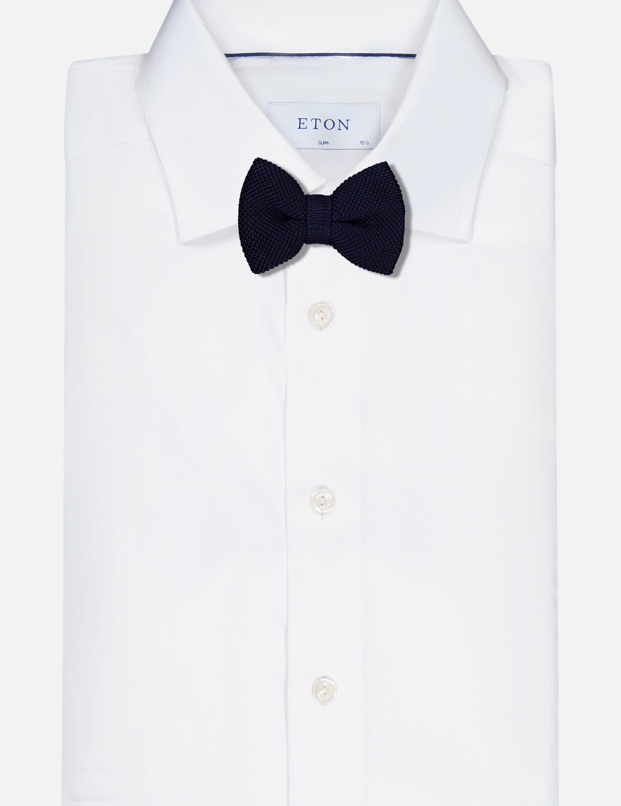 A folded white dress shirt with a classic collar is shown with a Ryan Silk Knit Pre-Tied Bow Tie by ROBERT TALBOTT in navy, neatly attached at the neck. The shirt features white buttons and an ETON brand tag inside the collar.