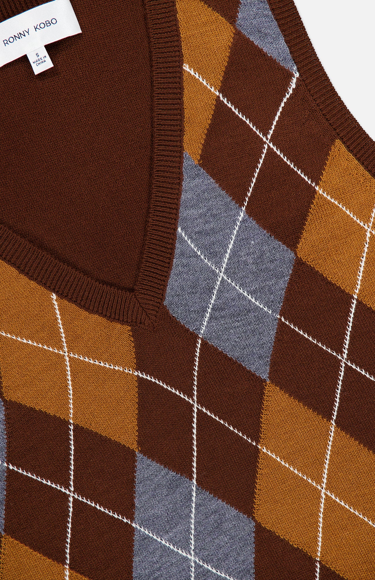 Close-up of the Gabriel Argyle Knit Vest by RONNY KOBO, showcasing a brown knit fabric with an argyle pattern in mustard and gray tones intersected by white lines. The vest features a V-neckline and ribbed edges for a timeless look, with a branded clothing label sewn onto the inner collar.