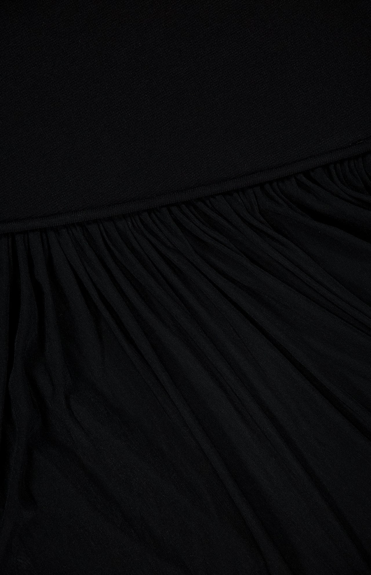 Close-up of the RONNY KOBO Meyer Draped Hemline Dress in black, featuring gathered pleats for a textured, wavy look. A smooth upper section contrasts with draped folds at the hem, creating a minimalist dress aesthetic.