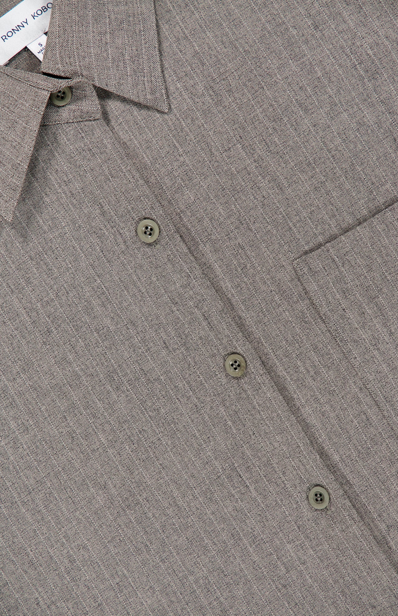 Close-up of the Niall Stripe Pocket Top by RONNY KOBO in grey, highlighting its pinstriped button-up design with a collar, three buttons, a pocket, and part of the label. The subtle vertical stripe pattern on the fabric makes this striped button-down blouse an ideal choice for versatile workwear.