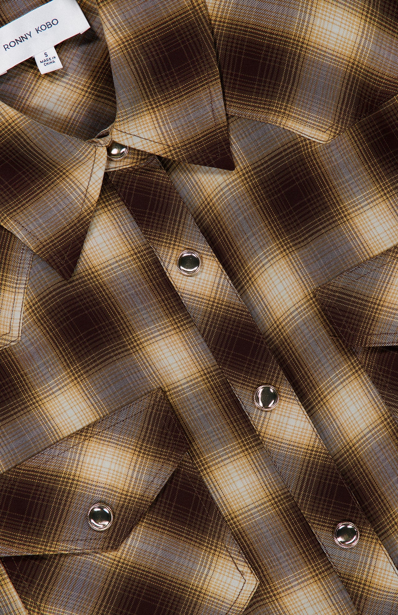 Close-up of the Tatie Plaid Western Shirt by RONNY KOBO, featuring a brown and beige plaid pattern, silver snap buttons, and two chest pockets. Perfect as a fall fashion piece, this western blouse includes a shirt tag in the corner that reads RONNY KOBO.