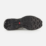 The image displays the black rubber outsole of the SALOMON XT-6 Shadow Sneaker, featuring a rugged traction pattern for enhanced durability and a small red triangular detail at the center.