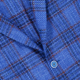 Closeup of the SARTORIO Wool Blend Plaid Sportcoat fabric in blue, showing a diagonal fold and two matching blue buttons. The material displays intersecting darker blue lines and fine orange stripes, highlighting the refined craftsmanship characteristic of this designer sportcoat.