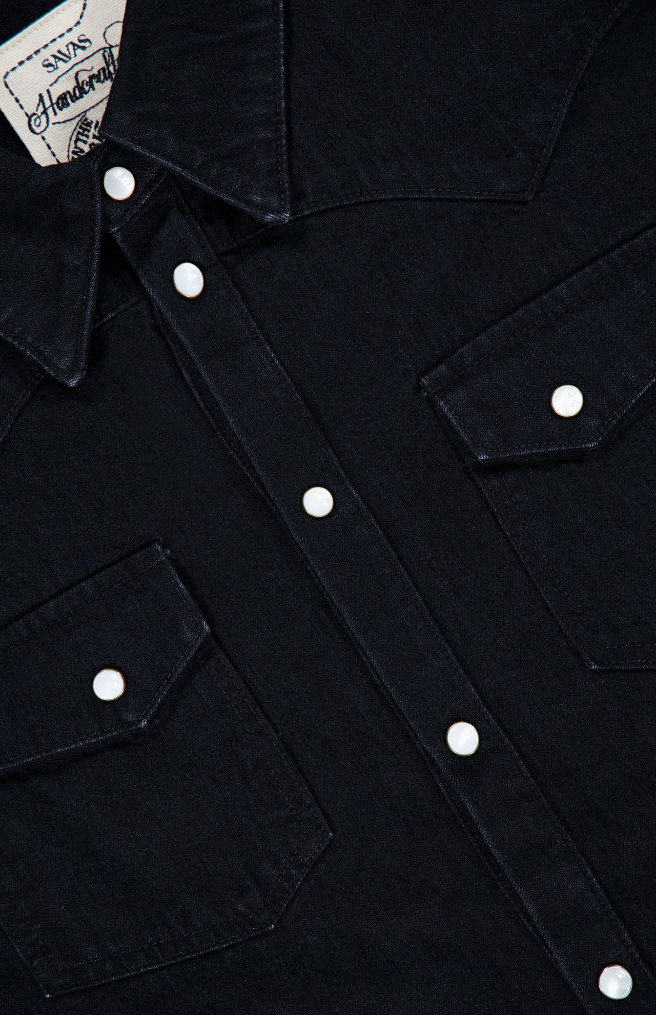 Close-up of the SAVAS Allen Denim Shirt in dark Japanese selvedge denim, featuring white snap buttons, a pointed collar, and two front chest pockets. The shirt tag is partially visible in the upper left corner.