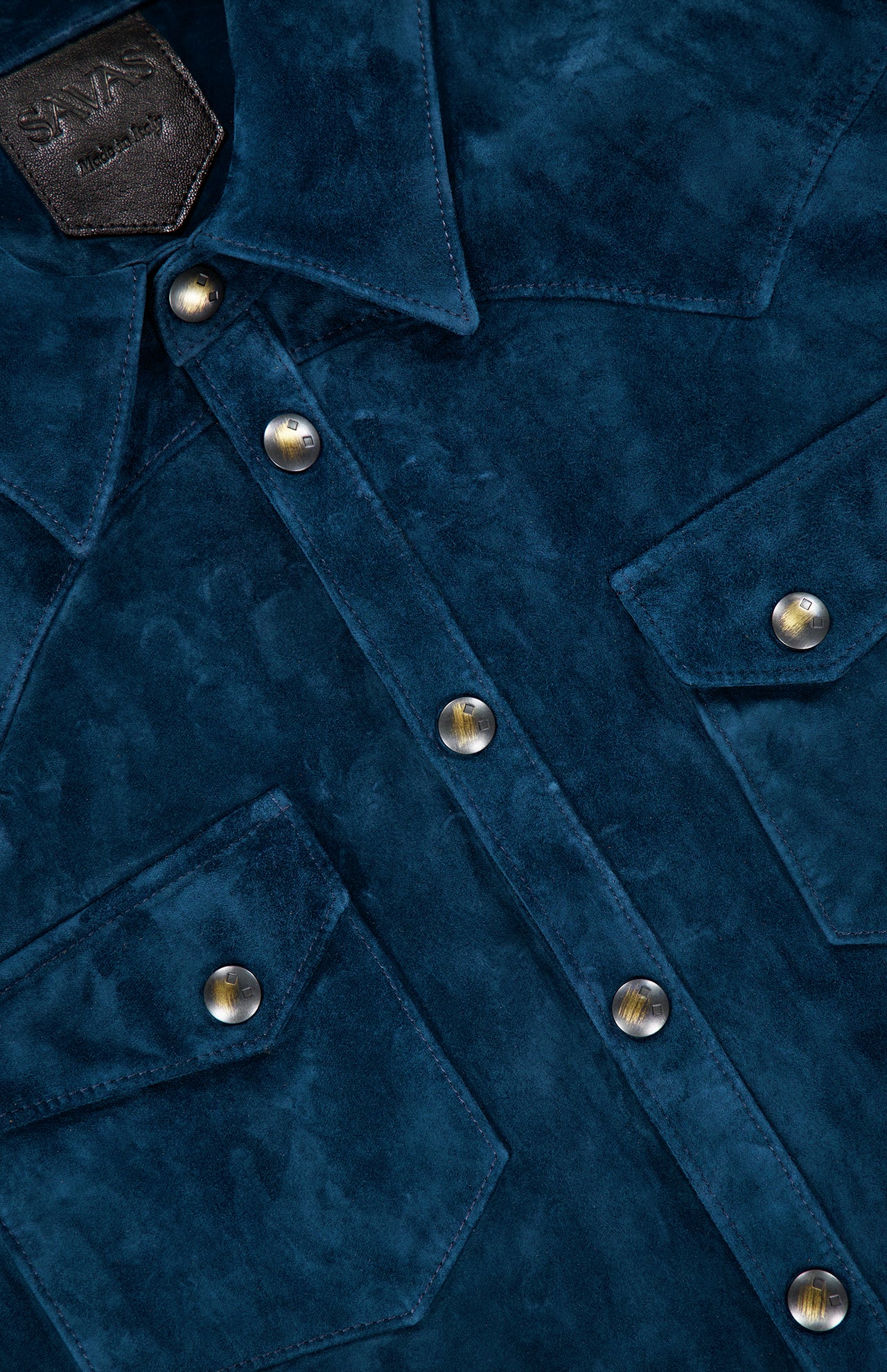 Close-up of the SAVAS Lowry Calf Suede Overshirt in blue, featuring metal snap buttons and two chest pockets. A partially visible leather tag with embossed text at the top left accentuates its refined Western-inspired style.