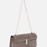 The Symmetry Chain Pouchette Bag by Savette is a taupe rectangular suede handbag accented with a silver chain strap and a sleek turnlock closure, shown against a white background.