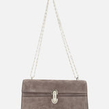The Symmetry Chain Pouchette Bag by Savette features taupe suede, a silver chain strap, and an elegant turnlock closure on the front, displayed against a white background.