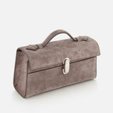 The Symmetry Pouchette Bag by Savette is a rectangular smokey grey suede handbag, crafted in Italy, featuring a top handle and a silver clasp, displayed against a plain white background.
