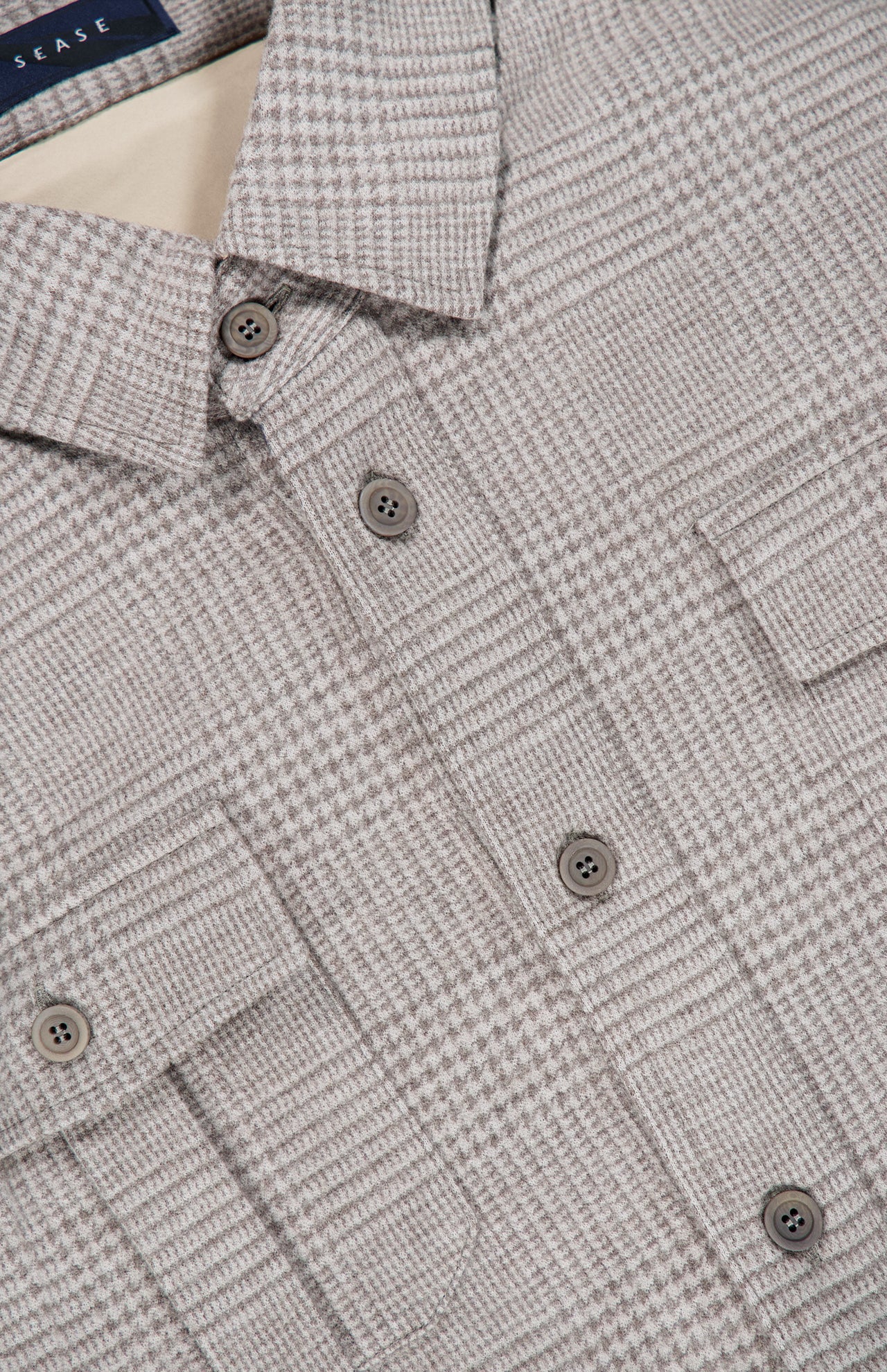 Close-up of the SEASE Glen Plaid Wool Overshirt, showcasing its light brown and beige glen plaid pattern with two chest pockets and plastic buttons. The collar and a portion of the label are visible at the top, emphasizing the refined wool texture inspired by the classic Prince of Wales check—making it an ideal option for business travel.