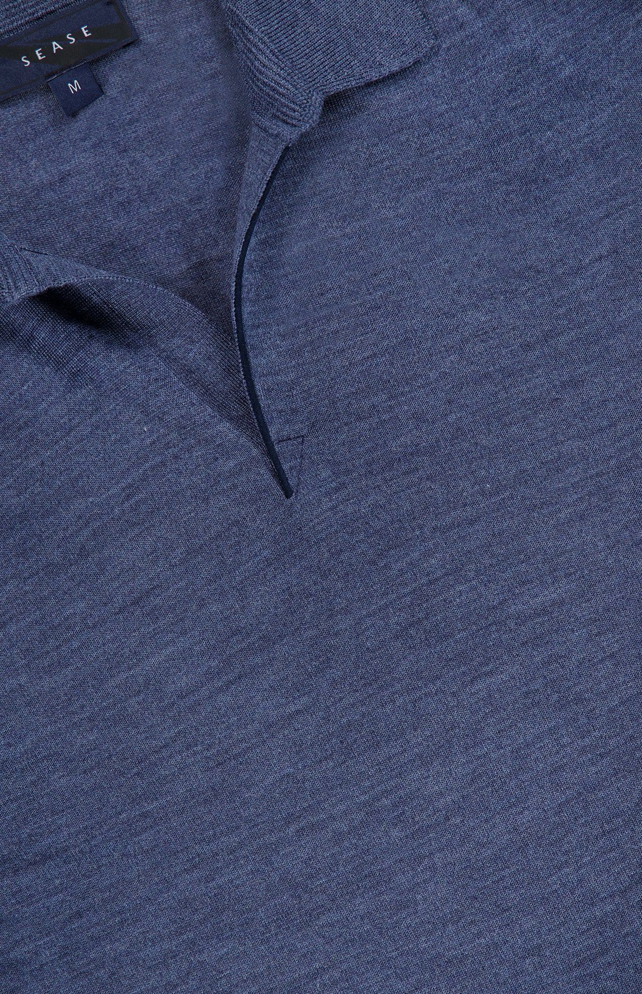 Close-up of the Lasca Long Sleeve Johnny Collar Polo Shirt in blue, showing a partial view of the collar and neck opening. Made from virgin wool, this SEASE piece features a black label with the brand name and size M visible at the top left.