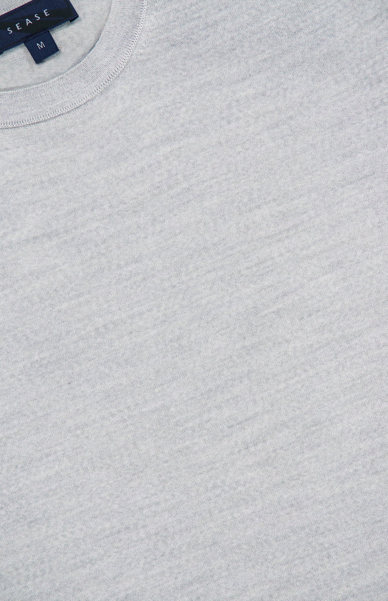 Close-up of the Lasca Long Sleeve Sweater by SEASE, crafted from luxurious Italian knitwear in light grey. This classic crewneck features a visible collar and a dark blue tag displaying the SEASE brand name and size M. The fabric exhibits a smooth, soft texture.