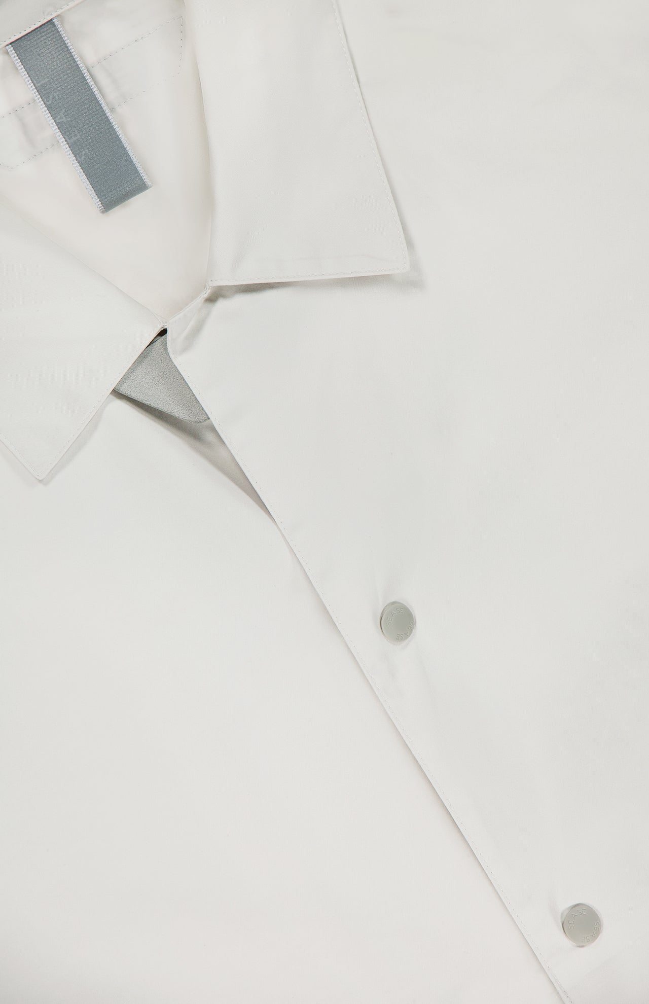 Close-up of the SEASE Spring Lifetime Drawcord Jacket, featuring a snap-front closure with silver snap buttons and a gray tag inside the collar, photographed against a white background.