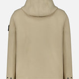 The Hooded Toggle Jacket by STONE ISLAND is presented in beige with a back view, showcasing long sleeves with buttoned cuffs and the signature Stone Island patch on the left sleeve. Made from a soft, fleece-like fabric, this versatile hooded jacket combines comfort with distinctive branding.