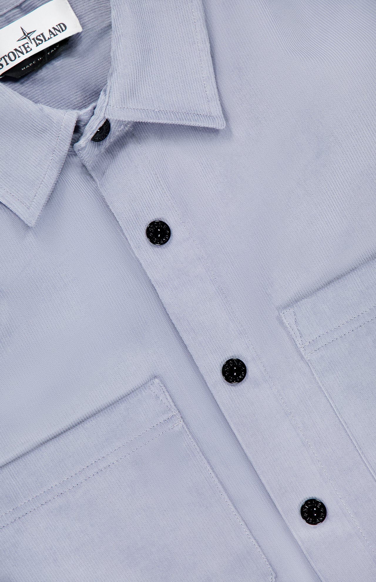 Close-up of the STONE ISLAND Long Sleeve Corduroy Shirt in light gray, featuring black buttons, two front pockets, and a branded label on the collar—an iconic men’s sportswear piece.