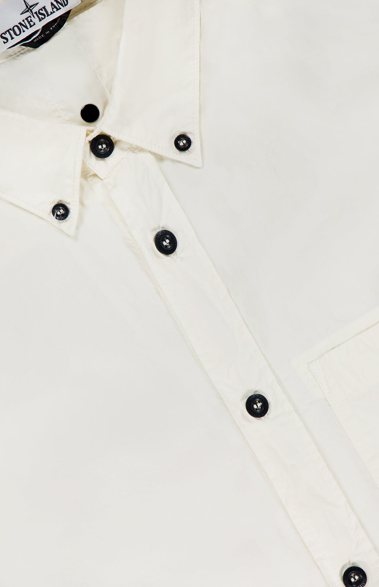 Close-up of a STONE ISLAND Long Sleeve Pocket Shirt in white, highlighting black buttons, a chest pocket, and a partially visible collar with branded label—capturing classic sportswear style.
