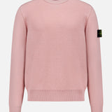 The STONE ISLAND Maglia Crewneck Sweater is a light pink, long-sleeve virgin wool top with ribbed cuffs and hem, and a black patch featuring the yellow-green circular logo on the left upper sleeve. Shown on a white background.
