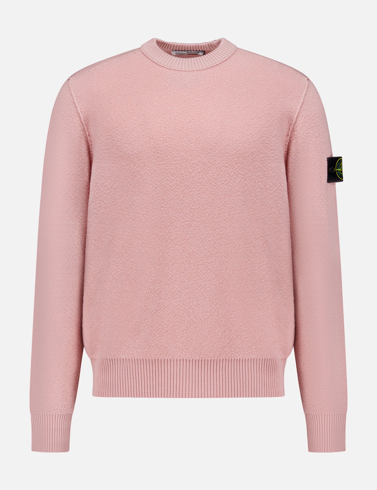 The STONE ISLAND Maglia Crewneck Sweater is a light pink, long-sleeve virgin wool top with ribbed cuffs and hem, and a black patch featuring the yellow-green circular logo on the left upper sleeve. Shown on a white background.