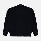 The Maglia RWS Cardigan by STONE ISLAND, crafted from luxurious virgin wool in black, is shown from the back against a plain white background. It features ribbed cuffs and hem and is distinguished by the iconic yellow and green STONE ISLAND patch on the left sleeve.