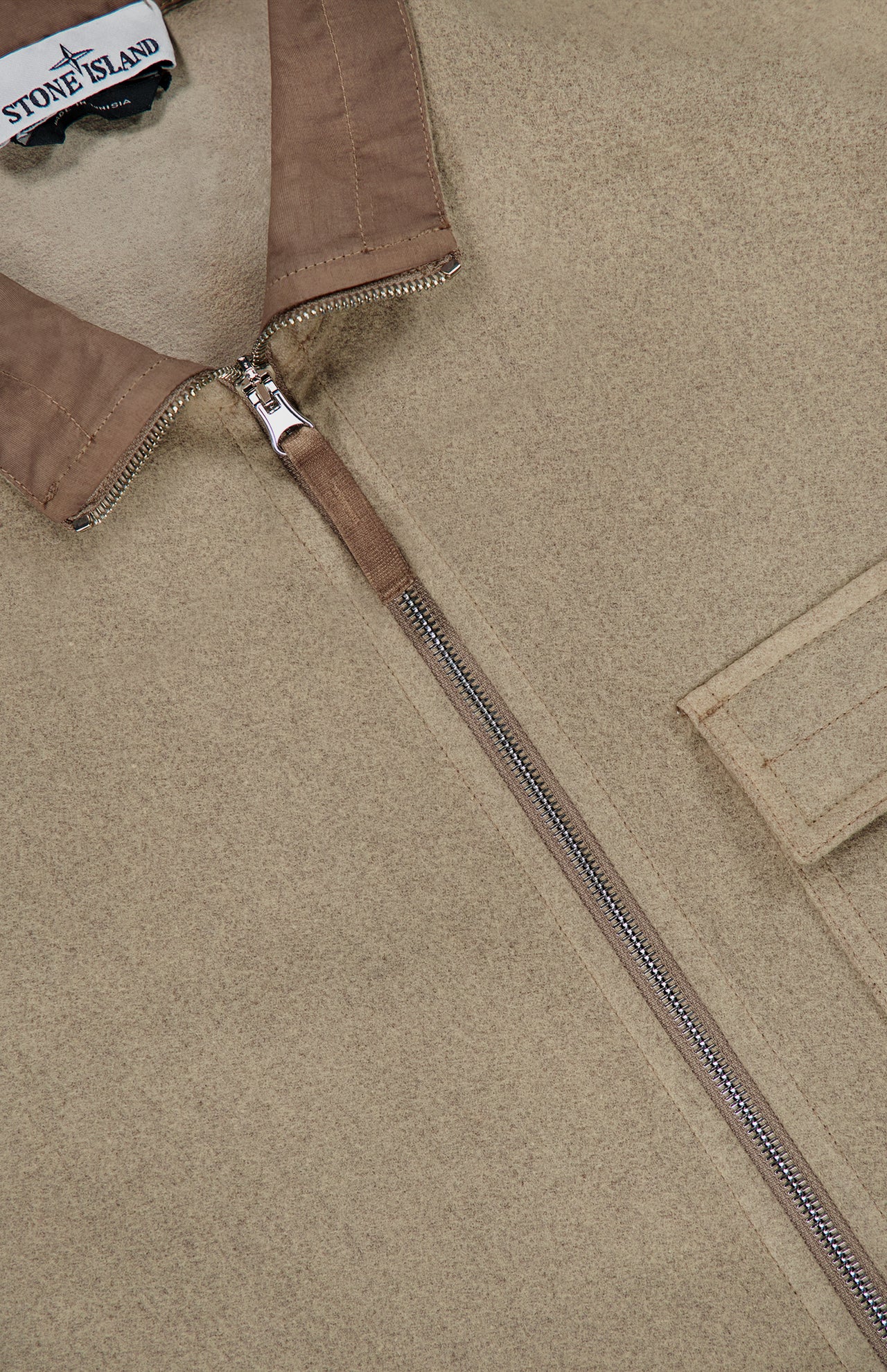 Close-up of the STONE ISLAND Stand Collar Zip Up Jacket in beige, a standout streetwear piece featuring a metallic zipper, flap pocket, and a fabric tag with the STONE ISLAND logo near the collar.