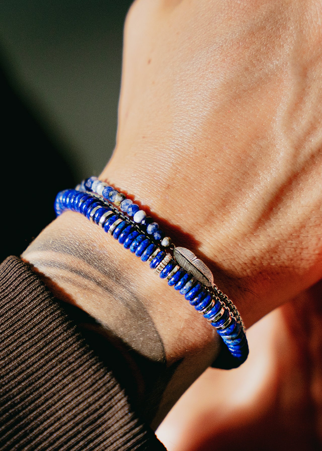 A close-up of a wrist wearing two stacked blue Ipanema Feather Bracelets by TATEOSSIAN—one with sodalite beads and one with disc-shaped beads—styled against brown clothing in natural light for a minimalist look.