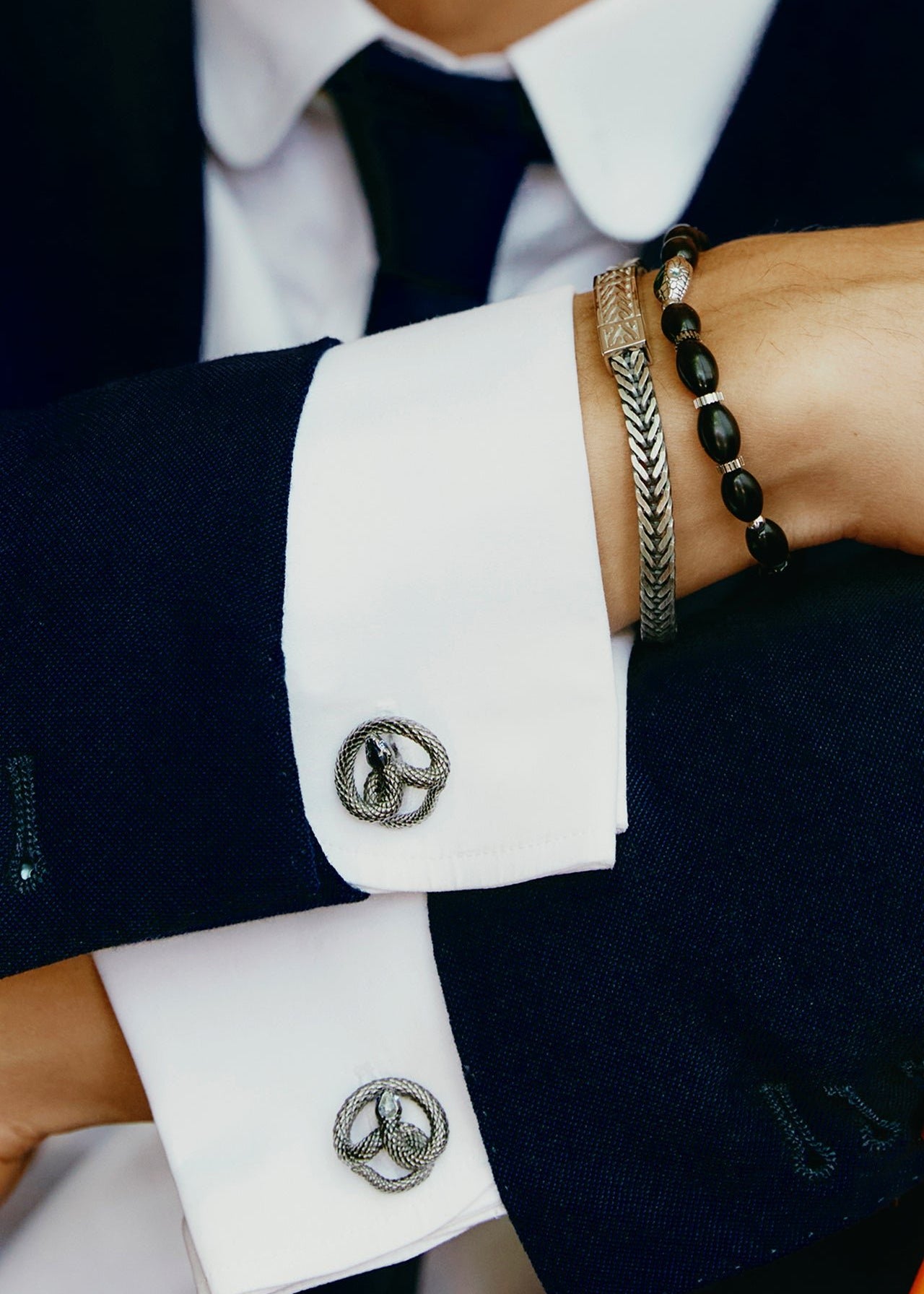A person in a dark blue suit and white shirt with decorative cufflinks and arms crossed, prominently displaying the TATEOSSIAN Viper Head Agate Bracelet to complement their polished formal look.