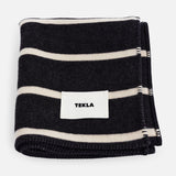 Folded dark charcoal and off-white striped TEKLA Striped Lambswool Blanket with a white brand label sewn on the front.