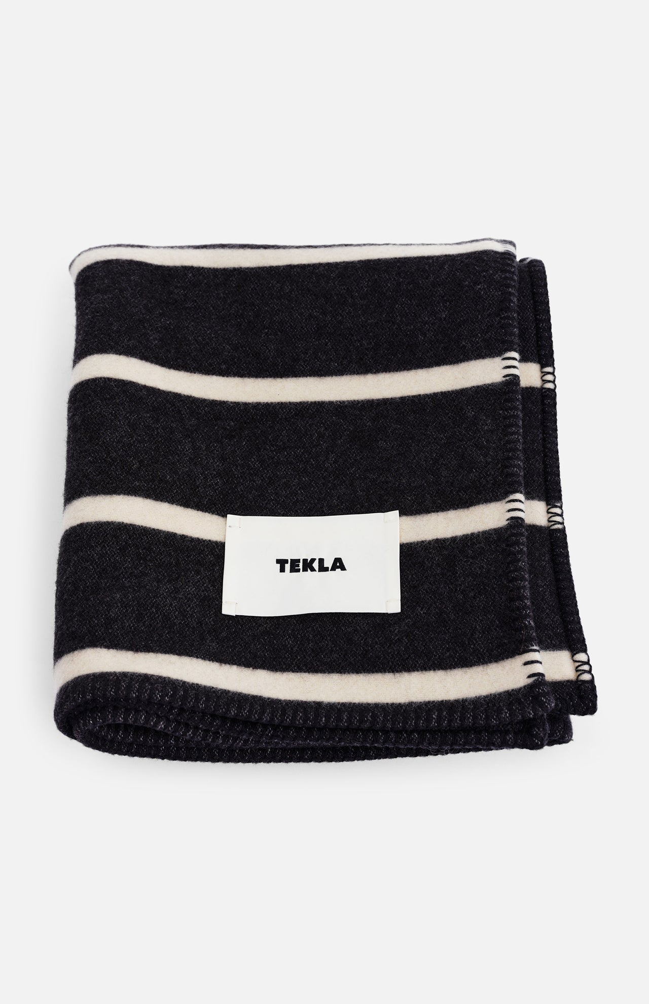Folded dark charcoal and off-white striped TEKLA Striped Lambswool Blanket with a white brand label sewn on the front.