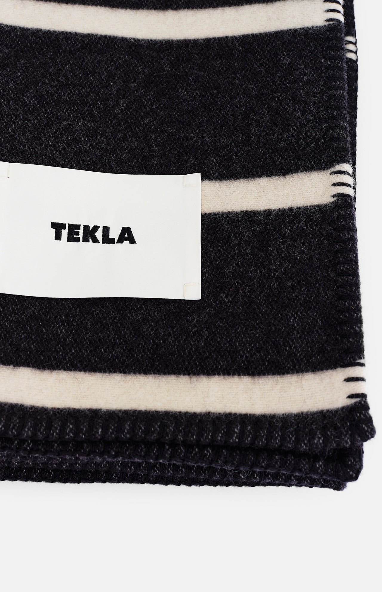 Folded black TEKLA Striped Lambswool Blanket with cream stripes and a white brand label.